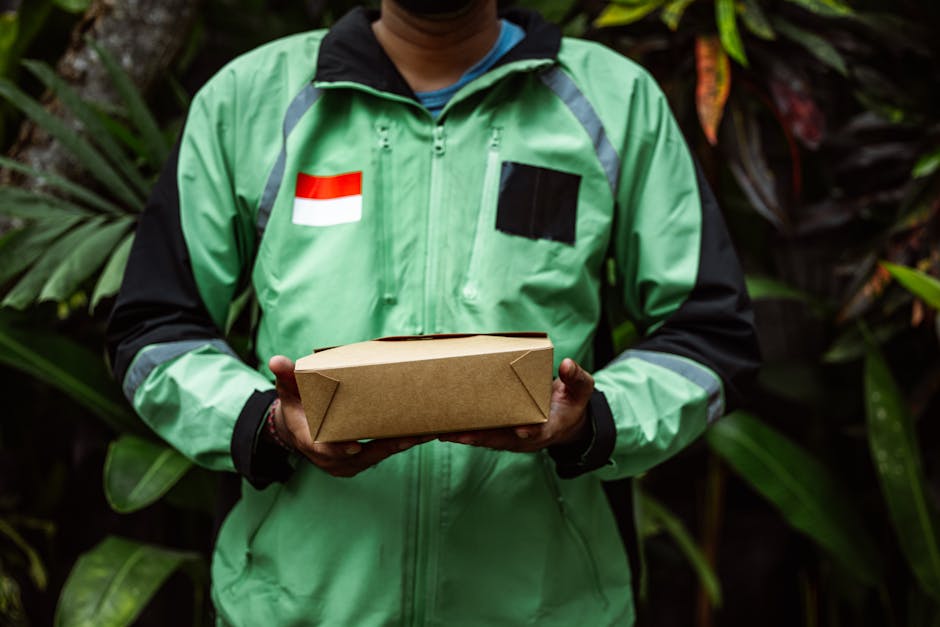 A delivery person in a green jacket with a red and white Indonesian flag patch and black accents holds a rectangular brown cardboard parcel with neatly folded edges, standing outdoors in front of lush