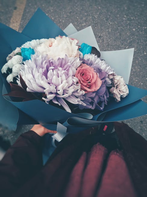 A bouquet of fresh flowers from Brent Flowers, featuring large white chrysanthemums, soft pink roses, purple spray roses, and small blue filler flowers, arranged together with vibrant green foliage. T