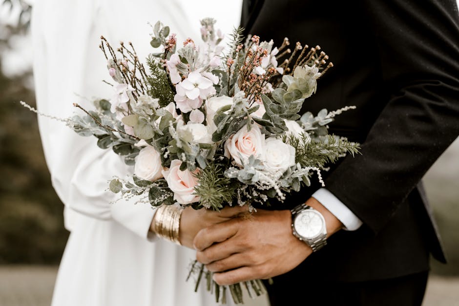 A close-up of a wedding couple holding a floral bouquet, featuring soft pink roses, white anemones, and pale pink orchids, accented with green eucalyptus and silvery dusty miller foliage. The bouquet