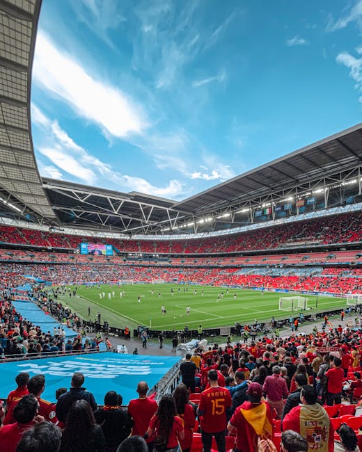 A wide view of Wembley Stadium during a football match, with bright blue sky and scattered white clouds overhead. The stadium is filled with spectators dressed in red and other colors, seated in tiere