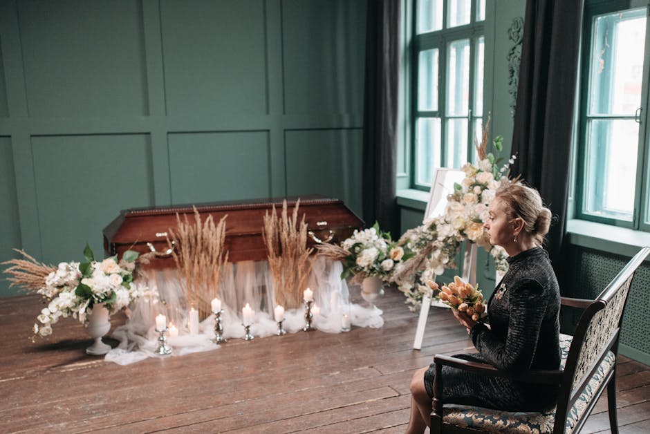 A woman dressed in black sitting on an ornate chair inside a room with teal paneled walls and large windows, holding a floral arrangement. In front of her is a long table decorated with white flower b