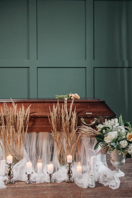 A floral arrangement consisting of white roses, peonies, and greenery in a white vase, placed on a wooden surface in front of a teal paneled wall. The display is complemented by sprigs of wheat and se